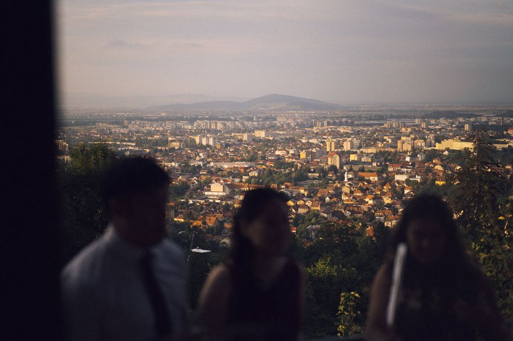 Blurred figures stand on a hilltop overlook with a panoramic cityscape and mountain backdrop at dusk.