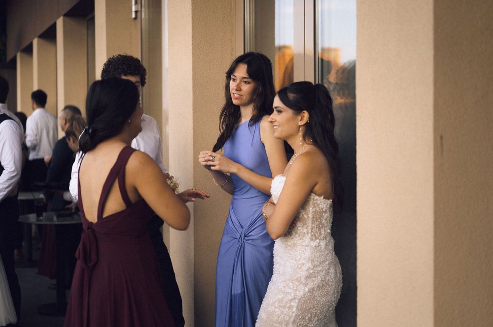 Bride in white lace gown converses with bridesmaids in blue and burgundy dresses at an outdoor wedding venue.