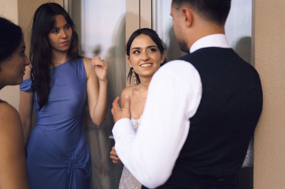 Bride in white dress smiles at groom in vest while bridesmaid in blue dress looks on at wedding.