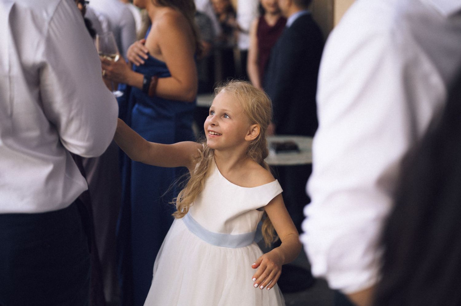 A young flower girl in a white dress with blue sash dances joyfully at a wedding reception.