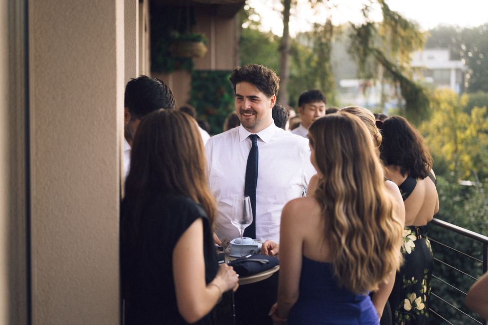 Guests socializing on an outdoor balcony at a formal event, man in white shirt and black tie holding a wine glass.