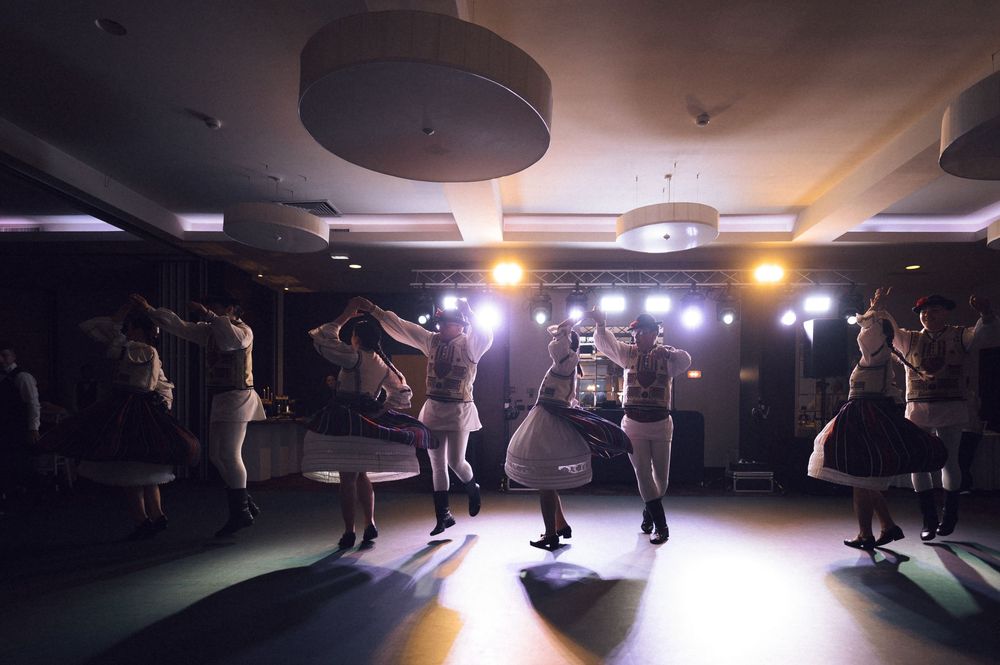 Group of dancers performing traditional folk dance in dimly lit ballroom with dramatic stage lighting.