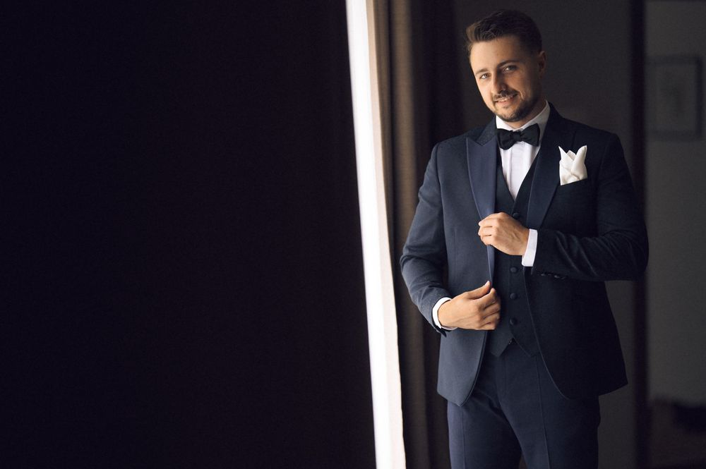 A well-dressed groom in a navy tuxedo with bow tie adjusts his jacket near a bright window.