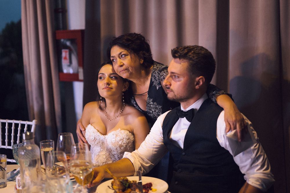 Bride and groom seated at wedding reception table with a woman embracing them from behind.