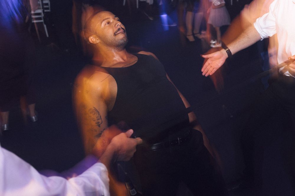 A muscular man in a black tank top dancing at a nightclub event, looking up with a smile.