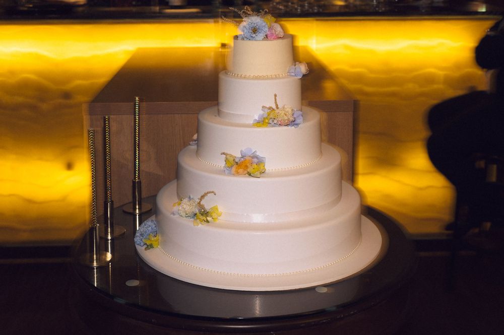 Elegant five-tier white wedding cake with delicate floral accents displayed on a stand against a warm golden backdrop.