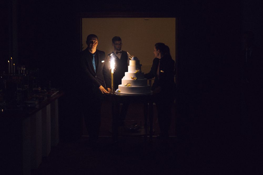 Three people gathered around a lit wedding cake in a dark elegant reception hall.