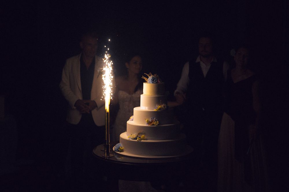 Couple cutting a white tiered wedding cake with sparklers in dark reception venue.