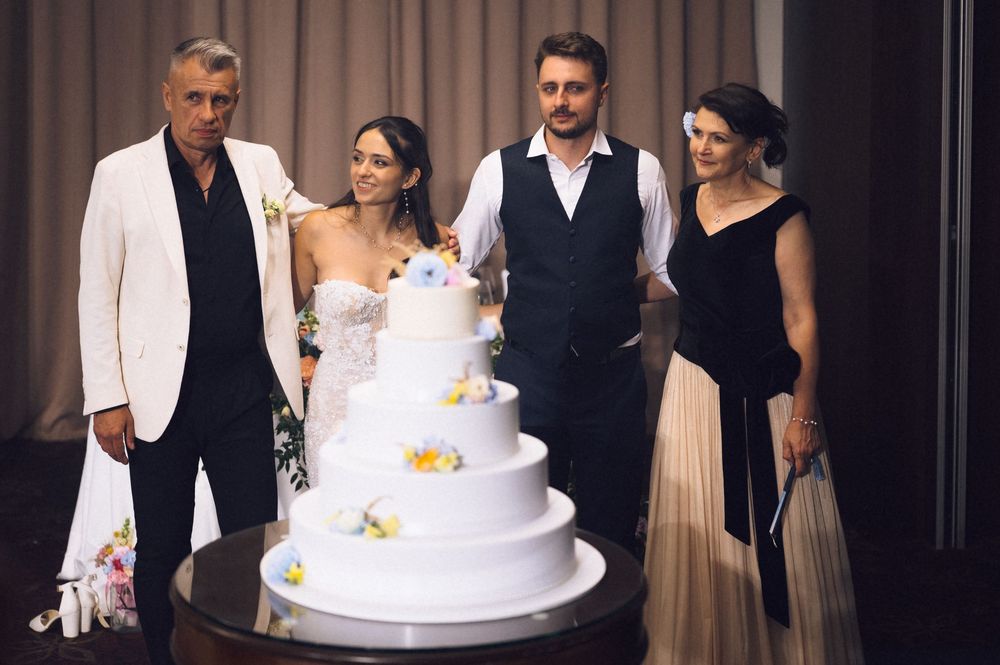 Four people posing together at a wedding reception beside a tall white tiered wedding cake decorated with colorful flowers.
