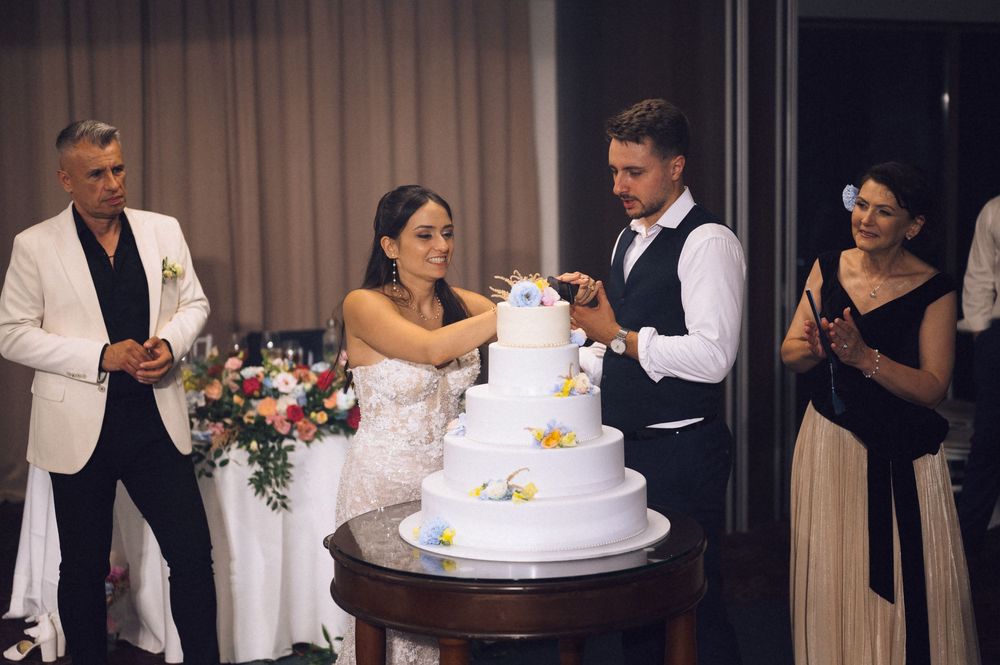 Bride and groom cutting a white four-tier wedding cake surrounded by guests at an elegant indoor reception.