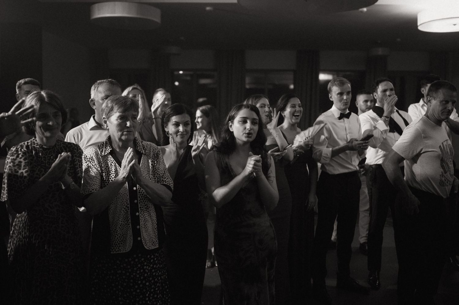 Black and white photo of guests clapping and dancing at an indoor evening event or wedding reception.