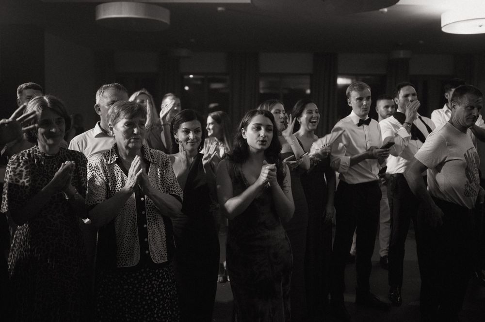 Black and white photo of guests clapping and dancing at an indoor evening event or wedding reception.