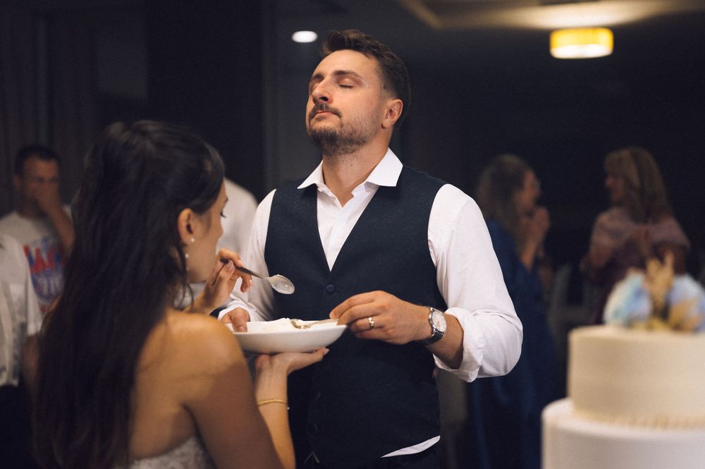 Groom in black vest and white shirt eating wedding cake at elegant indoor reception celebration.