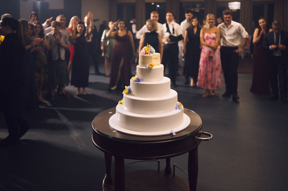 Elegant five-tier white wedding cake on wooden table surrounded by wedding guests at a reception venue.