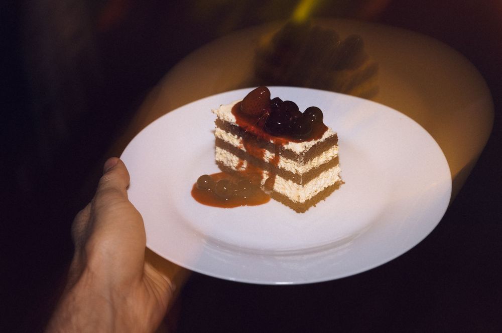 Elegant layered dessert with cream and chocolate toppings on a white plate, held by hand in dim lighting.