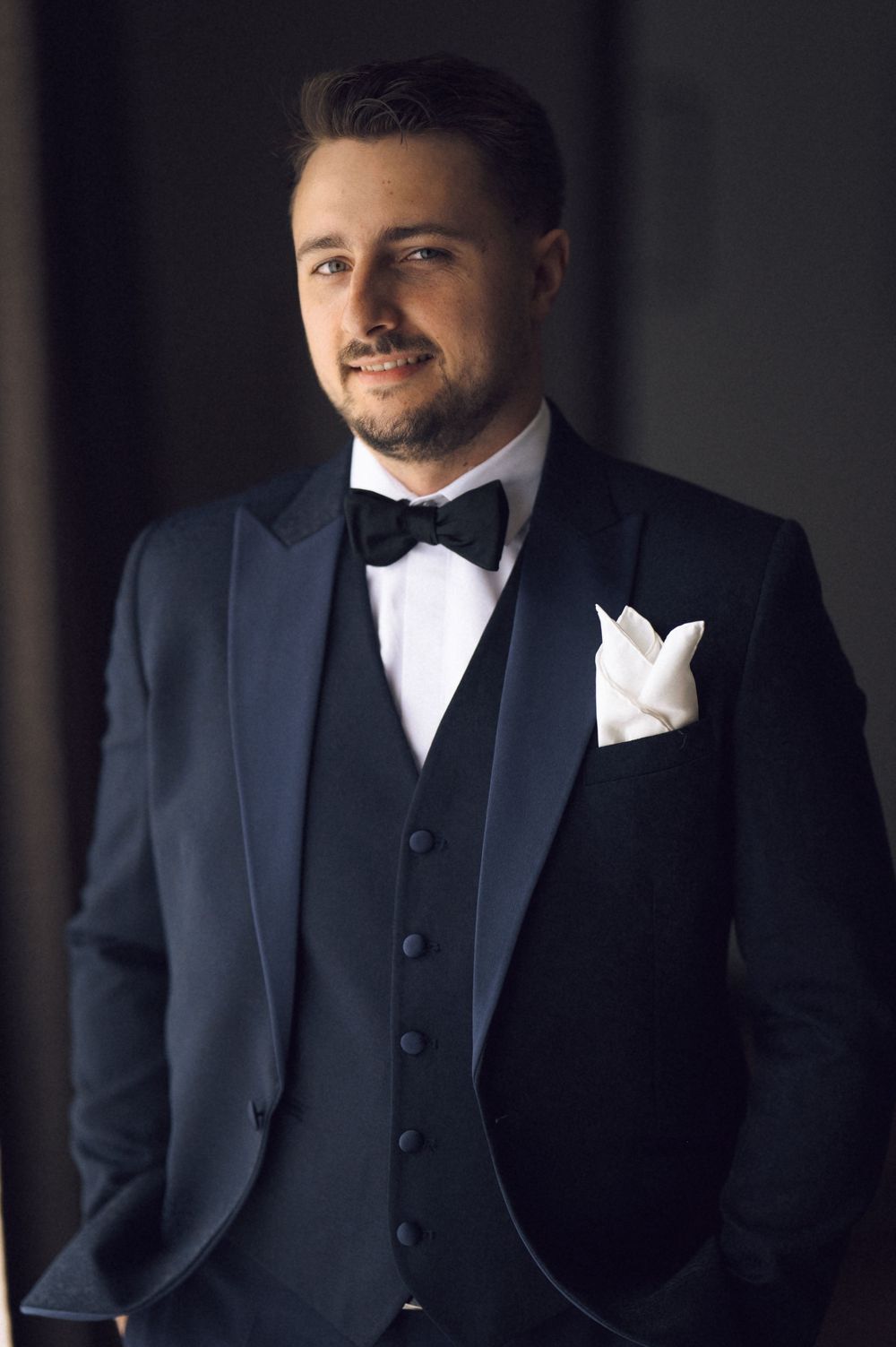 Elegant man in black tuxedo with bow tie and white pocket square posing confidently in a formal portrait.