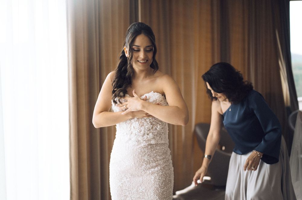 Bride in a strapless lace wedding gown being helped by bridesmaid near a window in a bright hotel room.