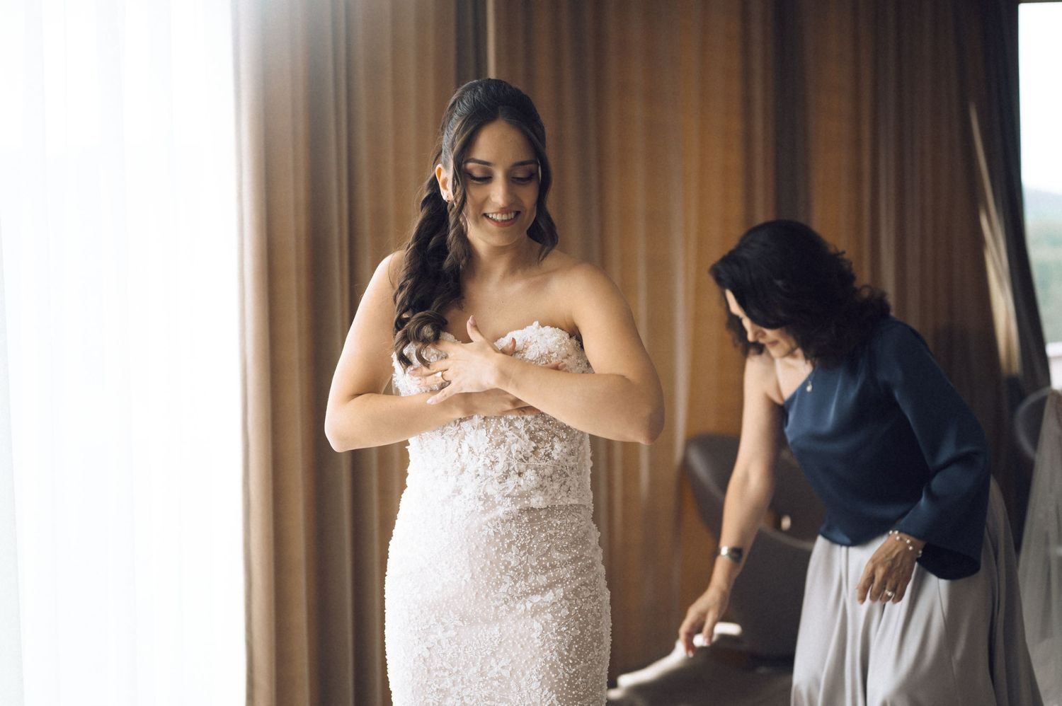 Bride in a strapless lace wedding gown being helped by bridesmaid near a window in a bright hotel room.
