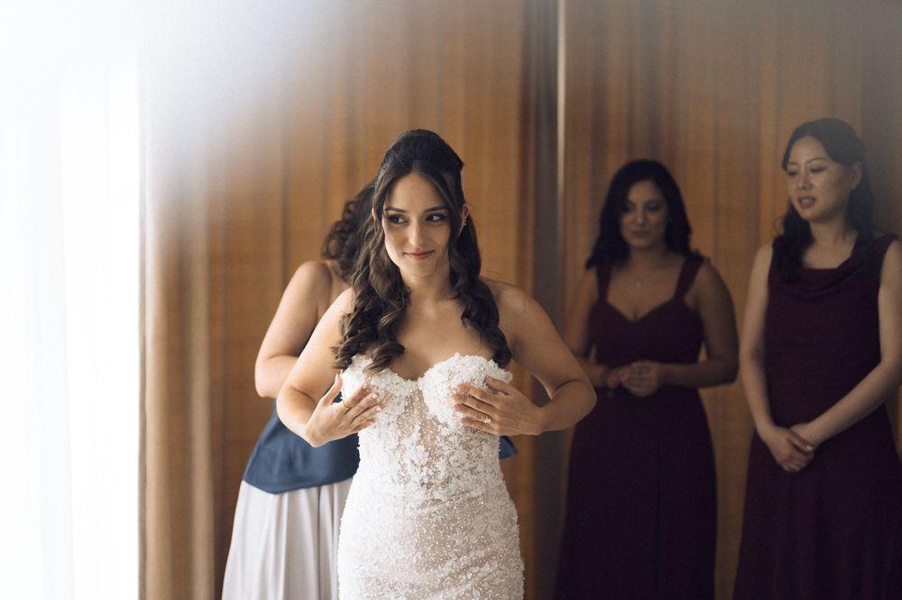 Bride in lace wedding gown being helped by bridesmaid while others in burgundy dresses look on.
