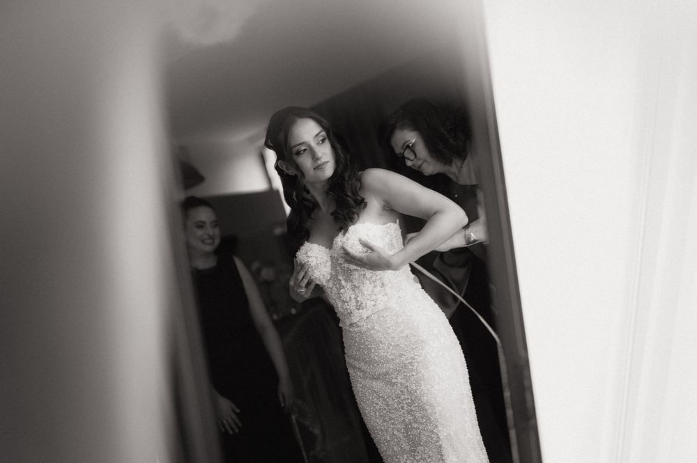 Black and white photo of a bride in a lace wedding gown being helped by bridesmaids while getting ready.