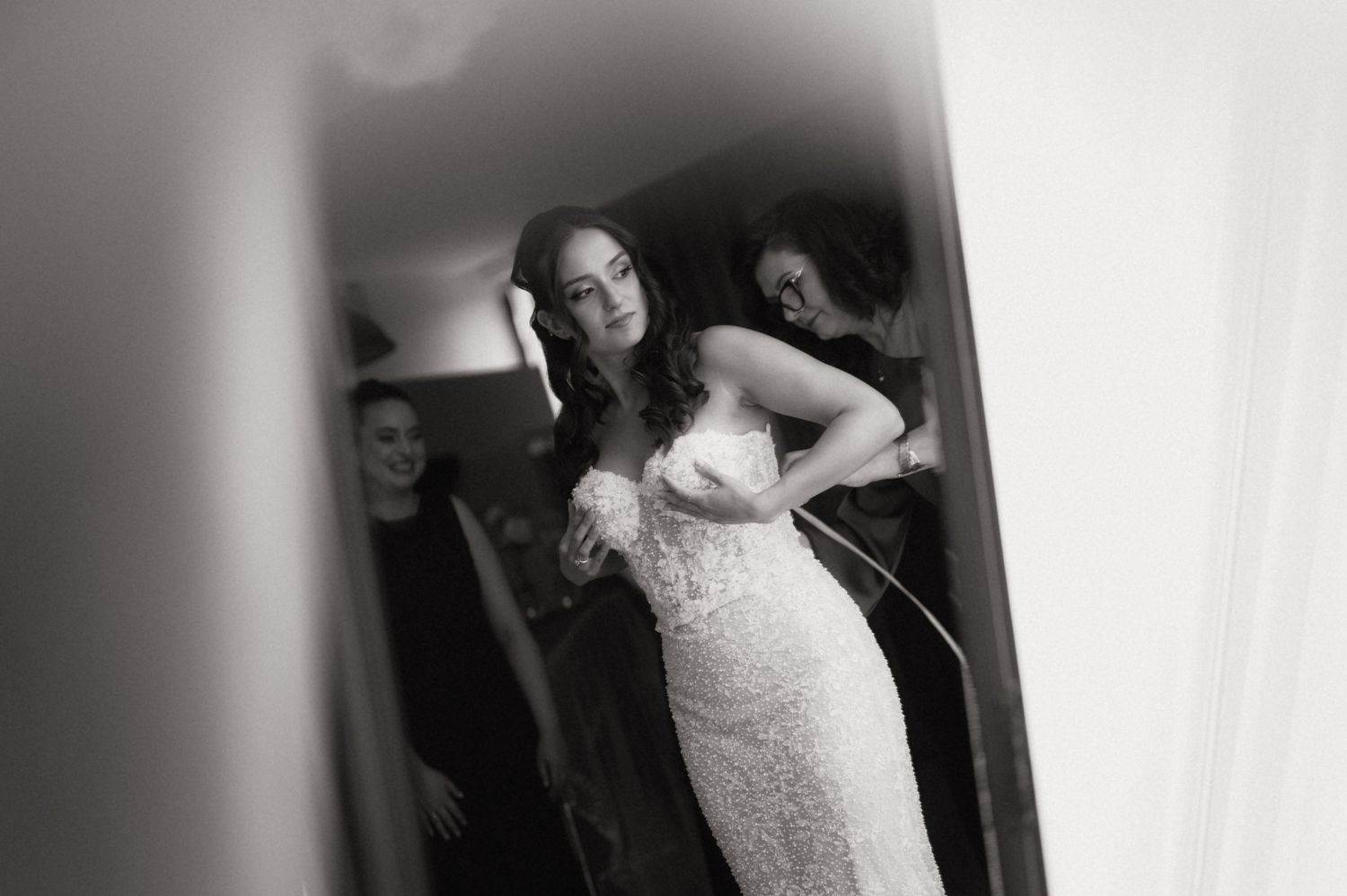 Black and white photo of a bride in a lace wedding gown being helped by bridesmaids while getting ready.