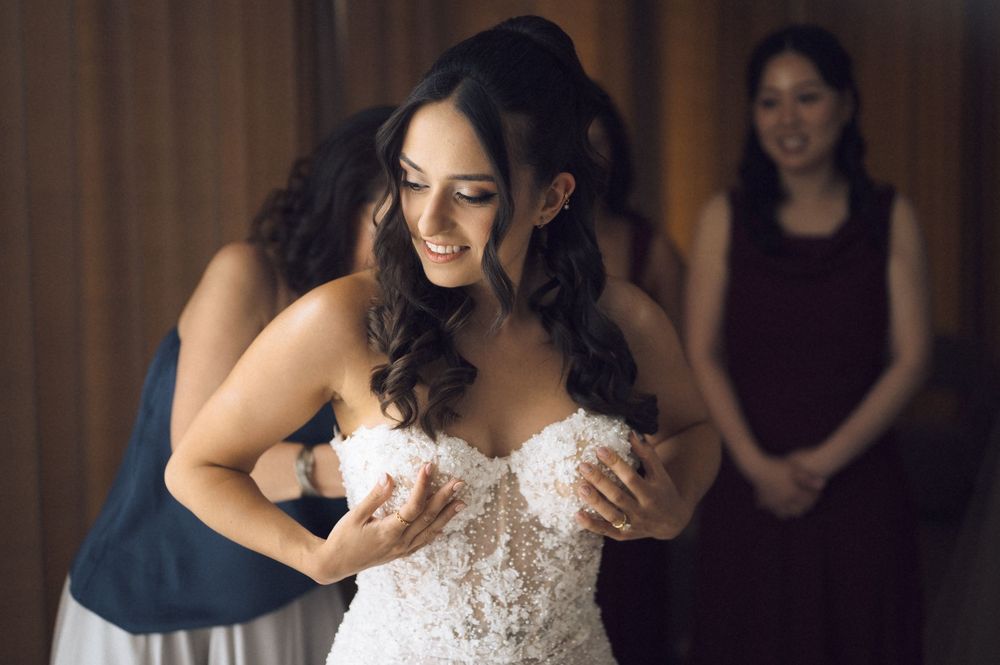 Bride smiling in lace wedding dress while bridesmaid helps her get ready, with another bridesmaid in burgundy watching.