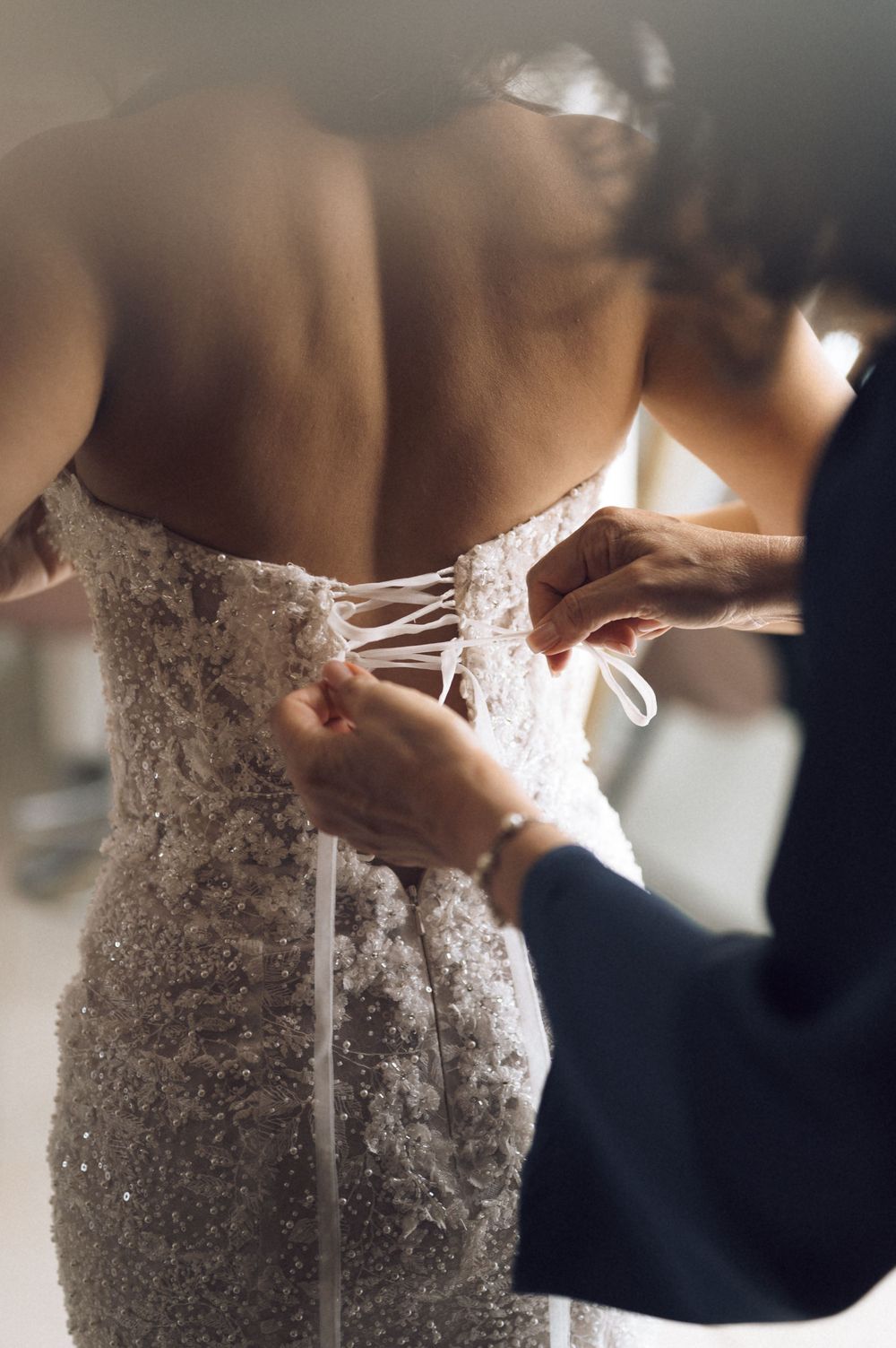 Someone lacing up the back of a beaded wedding gown, tying a white ribbon bow on a bride.