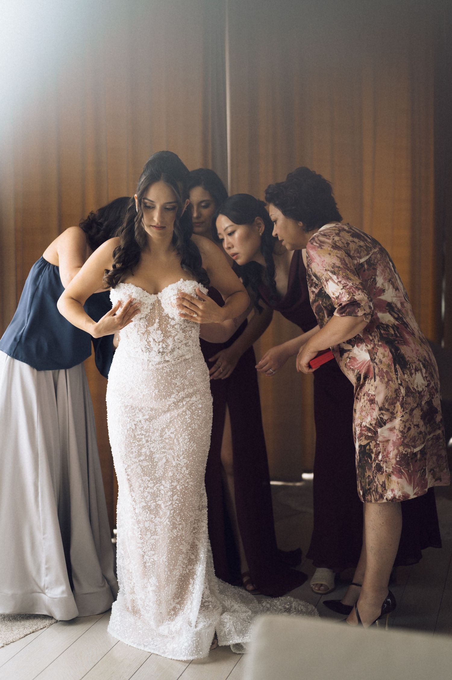 Bride in lace wedding gown being helped by bridesmaids and mother during wedding preparation.