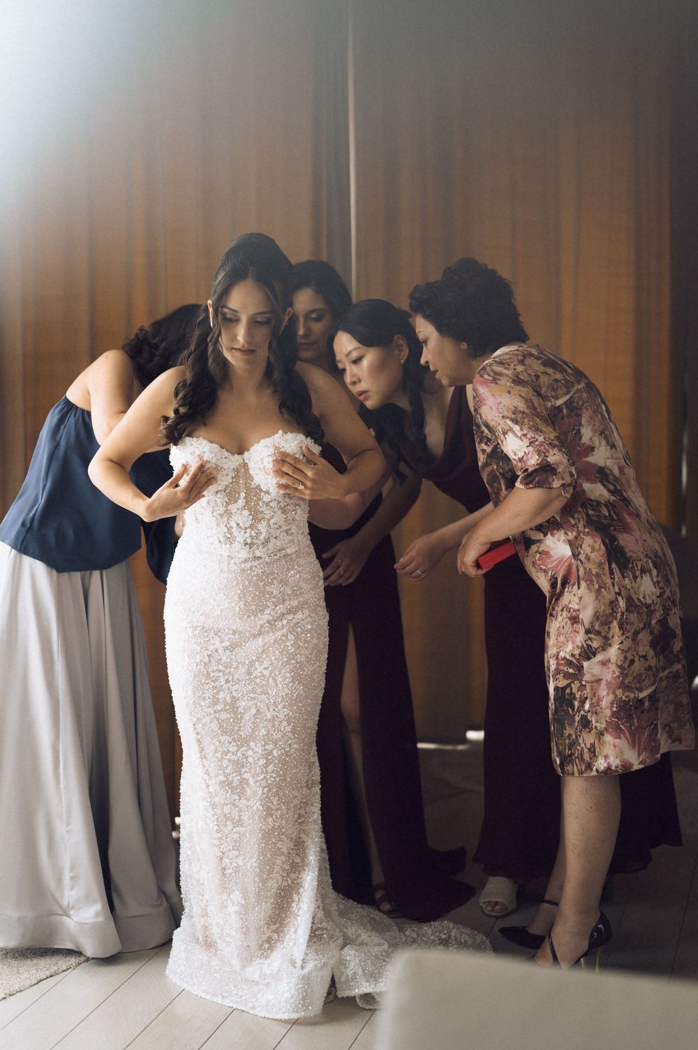 Bride in lace wedding gown being helped by bridesmaids and mother during wedding preparation.