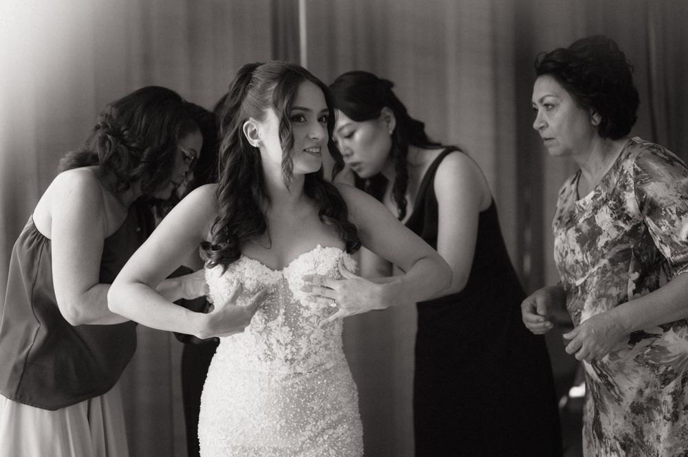 Bride in lace gown assisted by bridesmaids and mother while getting ready in black and white photo.