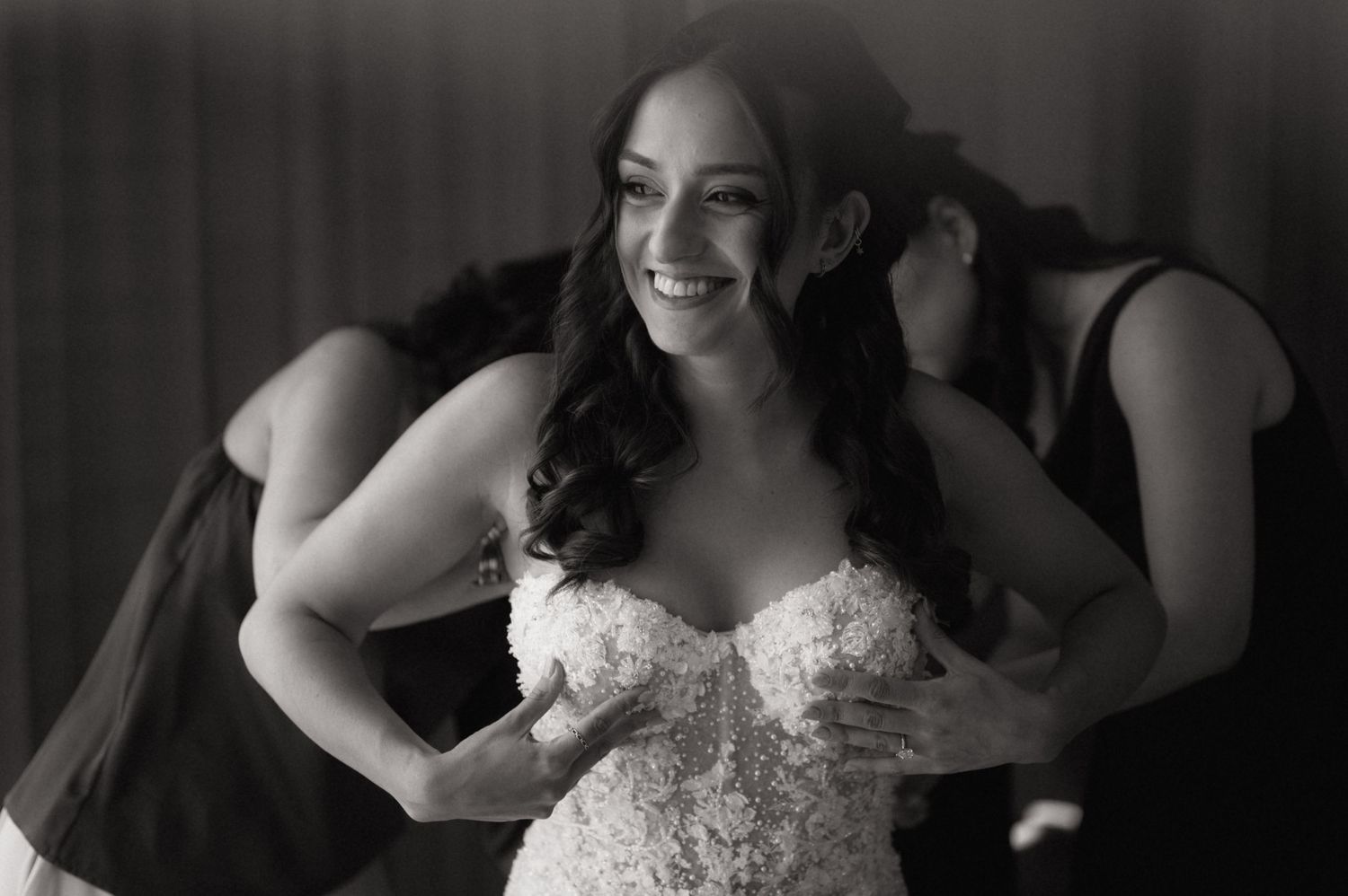 Black and white photo of a smiling bride in a lace wedding gown being helped by bridesmaids.
