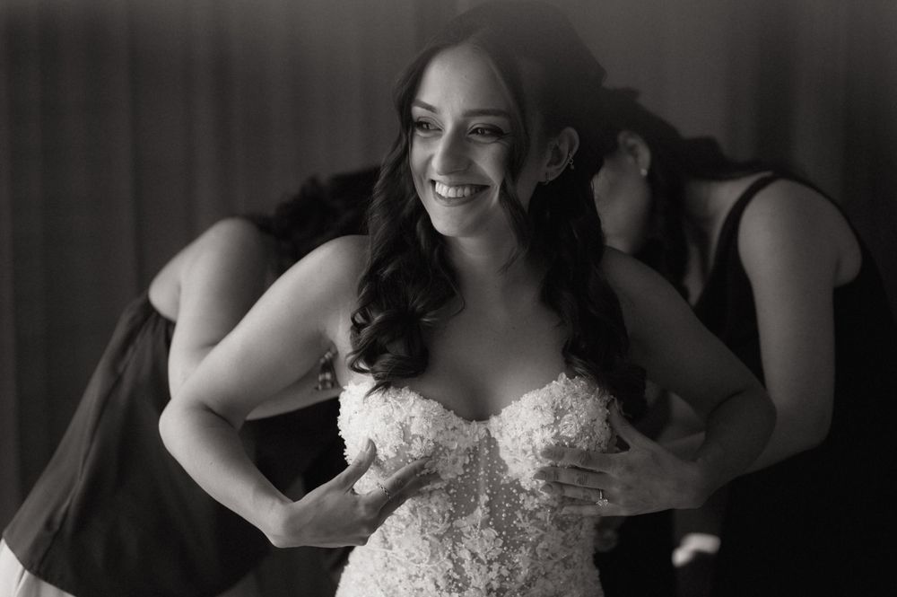 Black and white photo of a smiling bride in a lace wedding gown being helped by bridesmaids.