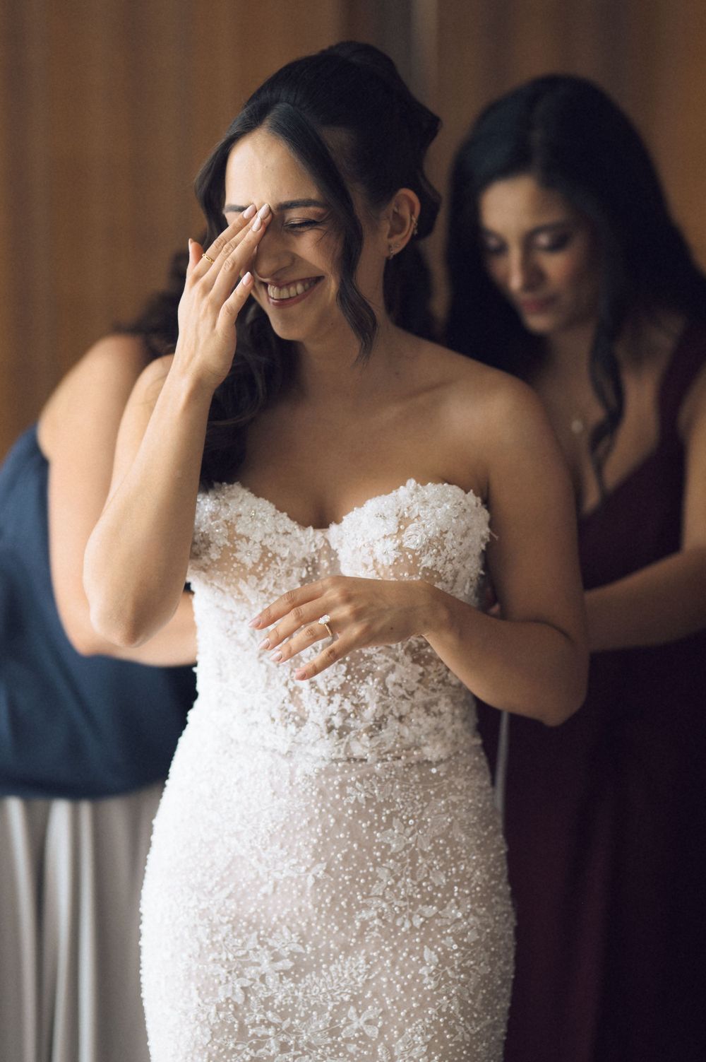 Emotional bride in strapless lace wedding gown wiping tears while bridesmaids help her get ready.