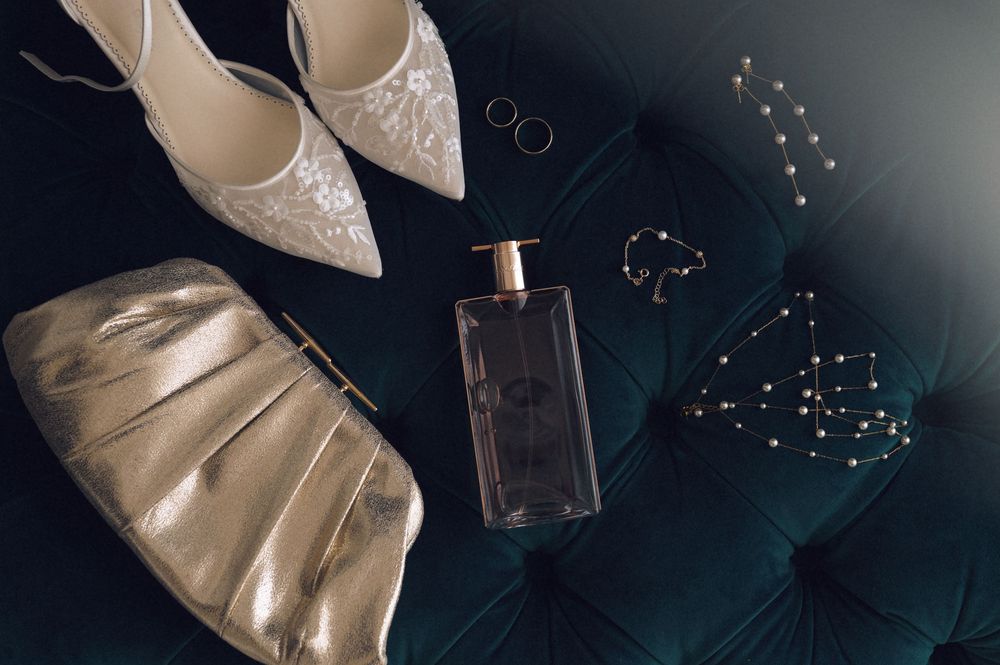 Elegant bridal flat lay with lace heels, gold clutch, perfume bottle, rings, and jeweled accessories on teal velvet.