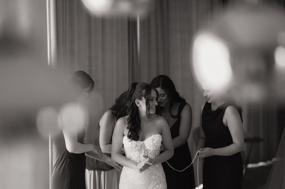 Black and white photo of a bride in a lace gown surrounded by bridesmaids helping her prepare on her wedding day.