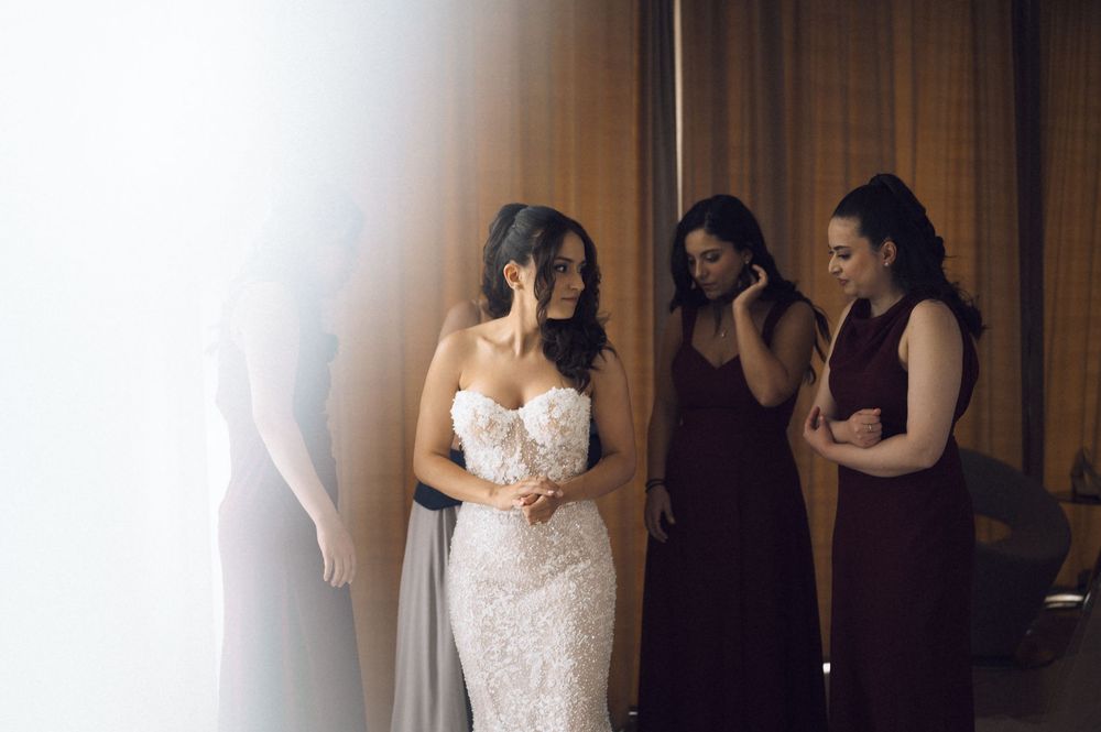 Bride in lace strapless gown stands with emotional bridesmaids in burgundy dresses during wedding preparations.