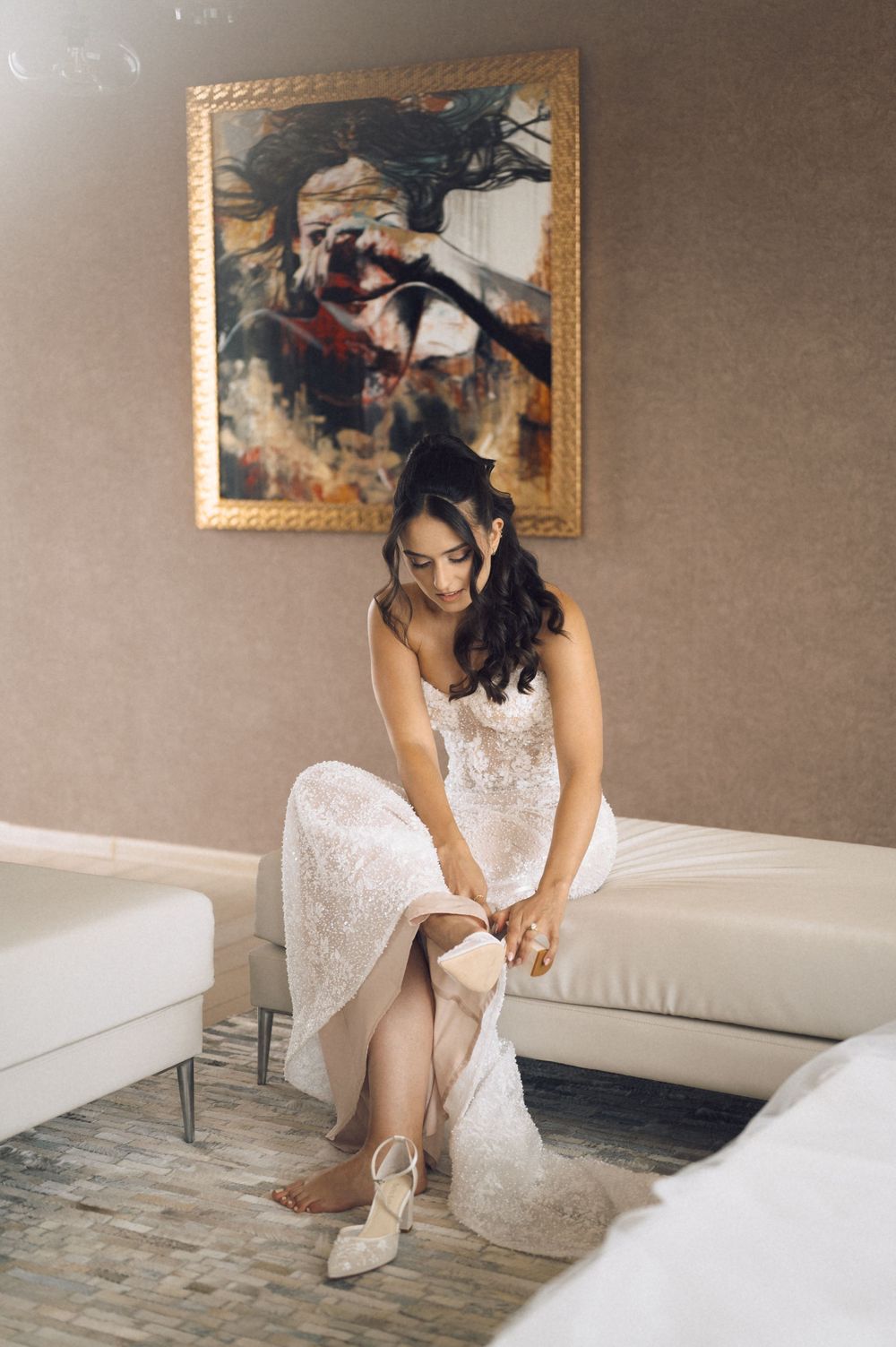 Bride in white lace gown sitting on sofa adjusting her heel shoe before wedding ceremony in elegant hotel room.