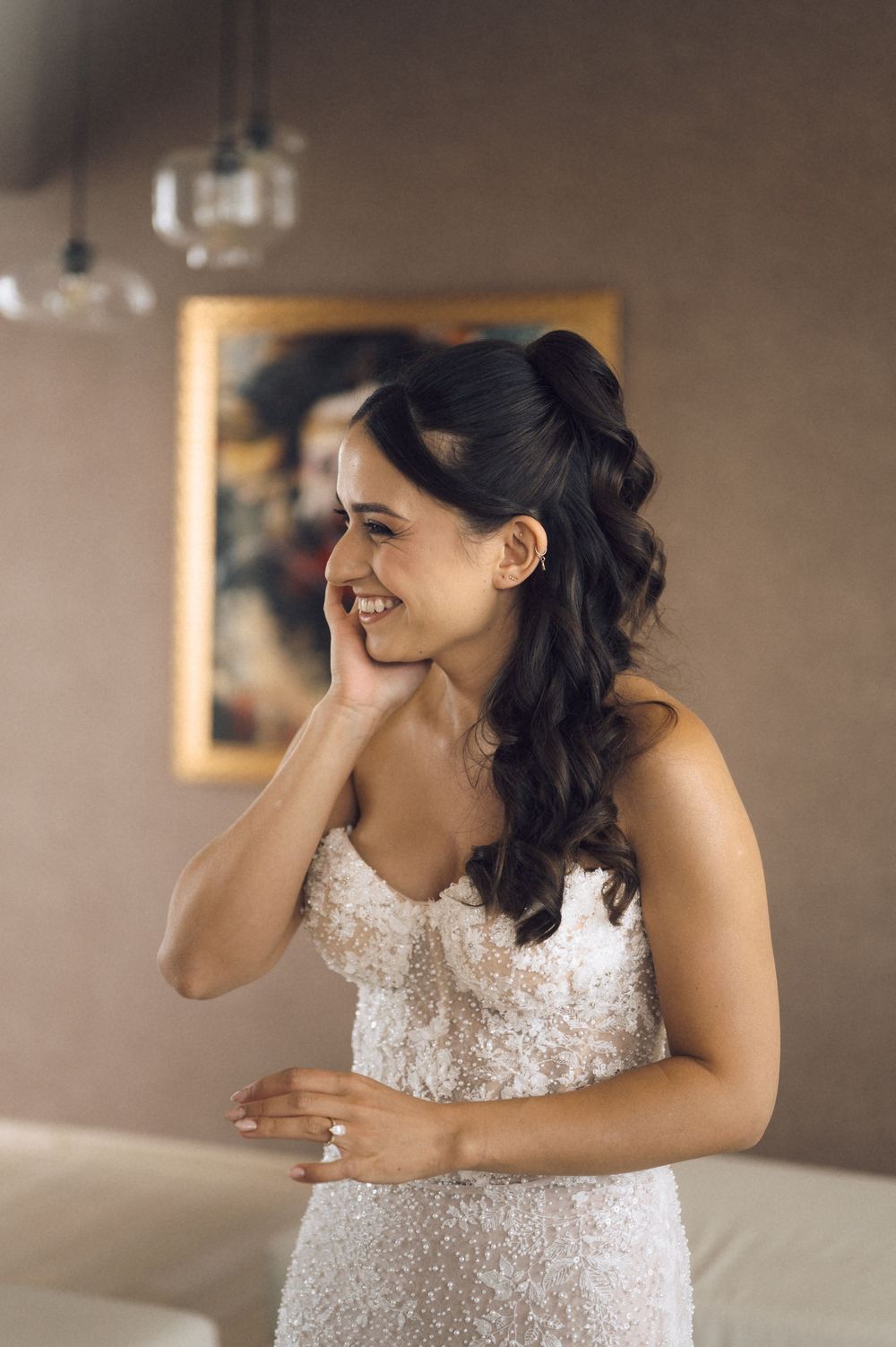Smiling bride in a beaded white wedding gown with half-up hairstyle, posing indoors near a framed painting.
