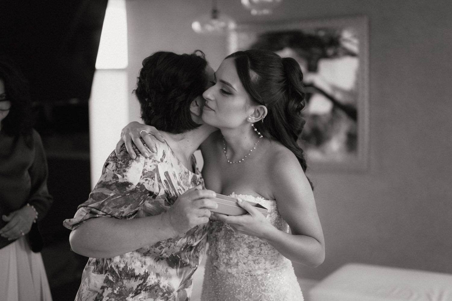 Black and white photo of a bride receiving a kiss on the cheek during wedding preparations.