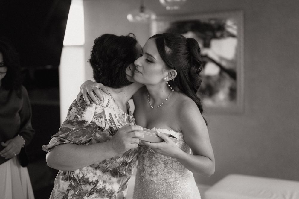 Black and white photo of a bride receiving a kiss on the cheek during wedding preparations.