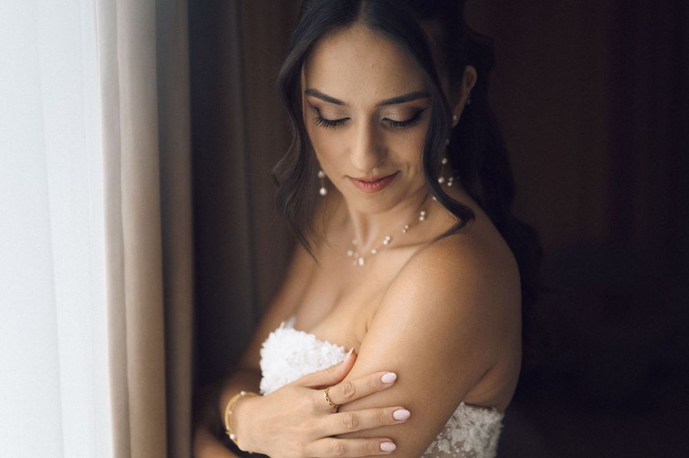 Elegant bride in white strapless gown with pearl necklace and earrings, looking down gracefully near a window.