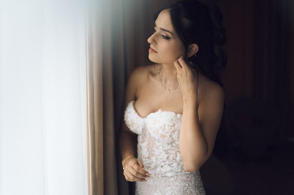 Bride in strapless lace wedding gown touching her necklace while gazing out a window in soft natural light.