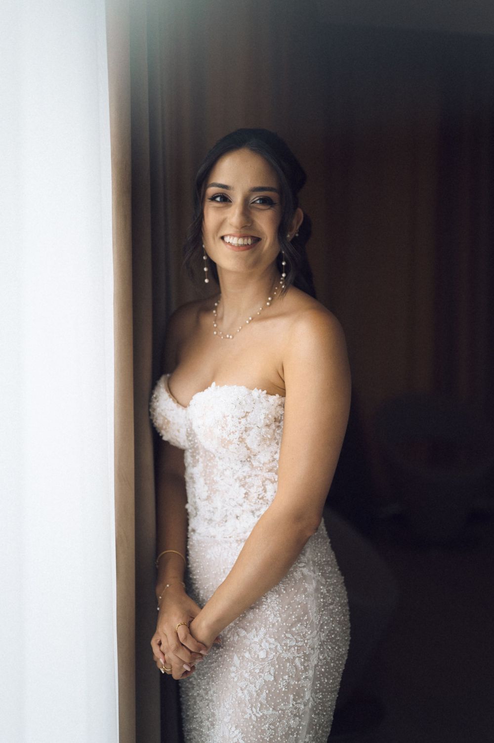 Smiling bride in strapless lace wedding gown with pearl necklace poses elegantly in soft natural light.