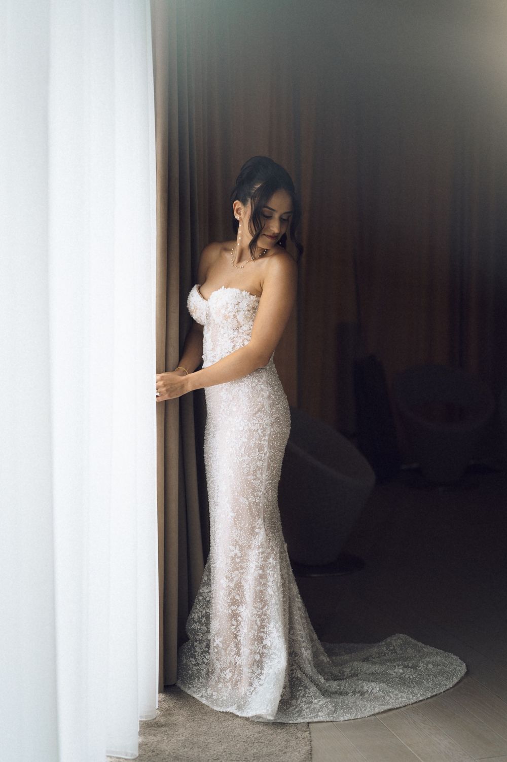 Bride in strapless lace mermaid wedding gown standing elegantly by sheer curtains in a softly lit room.