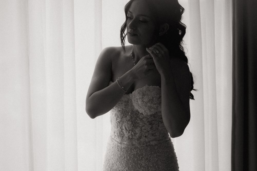 Black and white photo of a bride in a lace wedding gown adjusting her jewelry near a bright window.