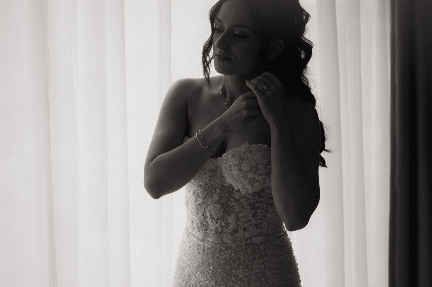 Black and white photo of a bride in a lace wedding gown adjusting her jewelry near a bright window.