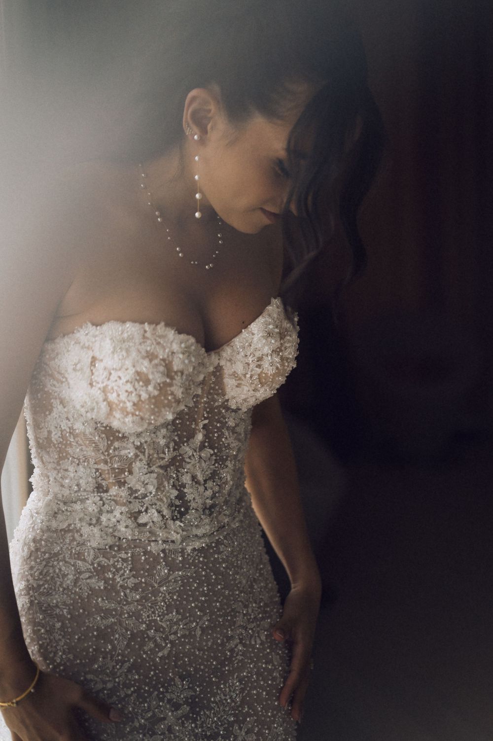 Bride in a strapless beaded lace wedding gown with pearl jewelry, posing in dramatic moody lighting.