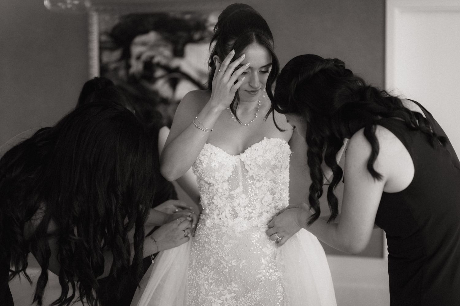 Black and white photo of a bride in a lace gown being comforted by two bridesmaids on her wedding day.