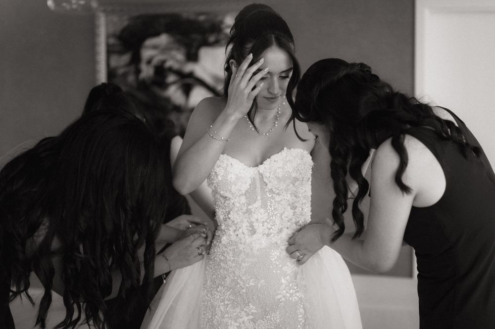 Black and white photo of a bride in a lace gown being comforted by two bridesmaids on her wedding day.