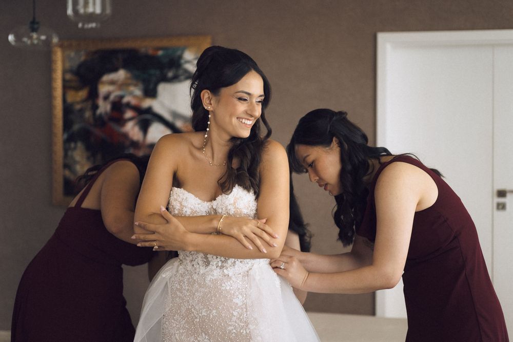 Bride in lace wedding gown smiles as two bridesmaids in burgundy dresses help her get dressed before the ceremony.